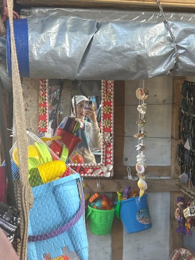 The image captures a selfie taken in a market stall in Bira, Indonesia. A person, wearing a white hijab and a light-colored sweater, is holding a mobile phone to take the photo, reflected in a decorative mirror. The mirror is framed with red and white decorations, incorporating shells and what appear to be small red and white flowers. In the foreground, a blue plastic bucket filled with colorful beach toys, including yellow and green shovels and red buckets, is visible, partially obscured by a woven bag. To the right of the mirror, a strand of seashells hangs as a decorative element. The stall's wooden walls are visible, with a tarp overhead. On the far right, sunglasses are displayed on a rack. A green bucket filled with more beach toys is also present. The scene suggests a beachside market setting in Bira.