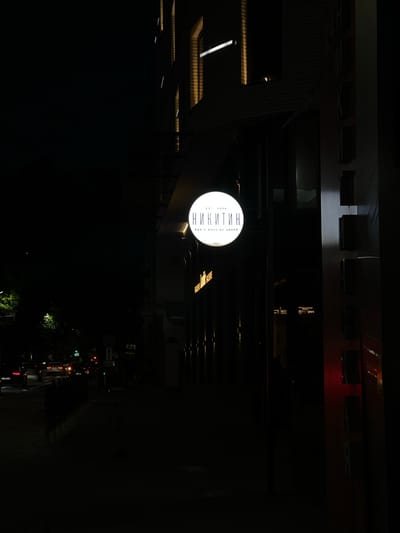The image shows a nighttime urban scene with a round, illuminated sign that reads "Никитин" in Cyrillic script. The sign also contains smaller text "EST 2024" and "ЕДА И ВИНО ВО ДВОРЕ" (Food and Wine in the Yard). The sign is attached to a building. The environment appears to be a city street at night with faint traffic lights and outlines of buildings visible. The scene is dark, with only a few light sources illuminating the sign and nearby structures. It is not possible to provide information about the city of Svkh Maslovskiy 1 Otdeleniye, Russia, since the only location indicator in the image is on the sign. There are no people visible.