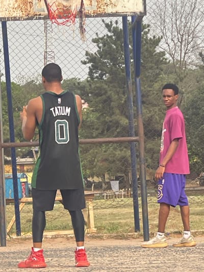 The image depicts an outdoor recreational area in Umunekwu, Nigeria. Two males are present on an unpaved, light brown earth surface. The male on the left is observed from behind, wearing a black and green basketball jersey with "TATUM" and "0" printed on the back, black shorts over black compression leggings, and red high-top athletic shoes. He is positioned facing a basketball hoop. The basketball hoop has a rusted metal backboard frame and a damaged red net, supported by blue poles. The male on the right faces towards the left, looking directly at the camera. He wears a magenta short-sleeved t-shirt, purple basketball shorts with a yellow "S" logo, a dark bracelet on his left wrist, and light-colored sneakers with "FILA" branded socks. A chain-link fence separates the foreground from the background, which features dense green trees and foliage. A distant communication tower is visible through the vegetation on the upper left. No direct interaction between the two individuals is evident.