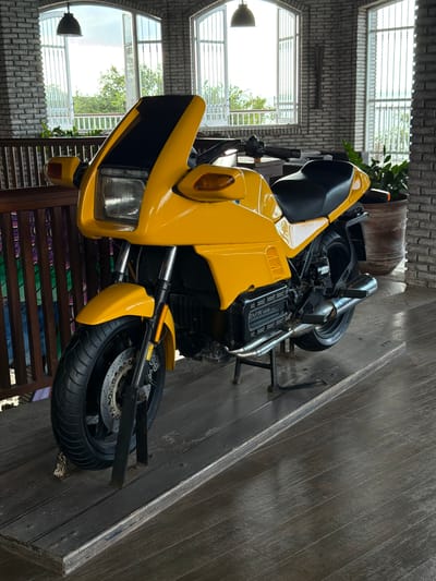 A bright yellow BMW motorcycle is parked on a wooden platform indoors, with large arched windows in the background offering a view of lush green trees and foliage. The motorcycle is the primary subject, facing left and slightly angled towards the viewer. It features a black seat, black handlebars, and a distinctive front fairing. The engine block is visible, with the "BMW" logo clearly marked. The motorcycle is supported by a black kickstand.
The environment appears to be a covered patio or restaurant area with exposed brick walls. The wooden flooring is rough-hewn planks. To the left of the motorcycle, a wooden railing with colorful vertical panels is visible. In the background, through the large windows, a bright, overcast sky is apparent, and the foliage suggests an outdoor natural setting. The scene is static and there are no people or animals present. The lighting is even, suggesting it's daytime with diffused natural light.