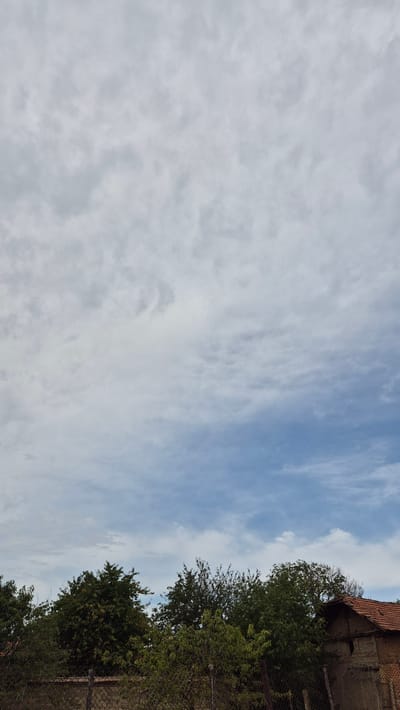 A view of the sky over Doirentsi, Bulgaria, captured from a ground-level perspective looking upwards. The sky occupies the majority of the frame, displaying a mix of overcast gray clouds with patches of blue sky visible towards the horizon. Below the sky, a line of dense green trees forms a dark silhouette against the lighter sky. To the right, a portion of a small, rustic building with a terracotta-tiled roof is visible, constructed from brick and mud. In the foreground, a chain-link fence runs horizontally across the lower part of the image, separating the viewer from the trees and building. The scene depicts a rural or semi-rural environment under a cloudy sky.