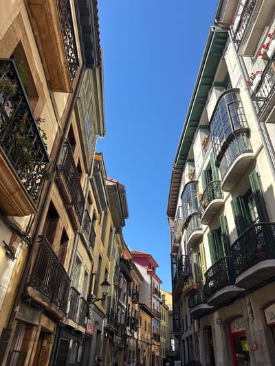 This image captures a narrow street in Oviedo, Spain, with tall, narrow buildings on either side, leaning towards each other and creating a canyon-like effect. The architecture is characteristic of European old towns, featuring stone facades, wrought-iron balconies adorned with potted plants and flowers, and large, arched windows. The buildings vary in color, with hues of beige, ochre, yellow, and white, punctuated by green shutters on some windows. The sky above is a clear, vibrant blue, indicating a sunny day. The street itself appears to be paved with stone. Some of the buildings have exposed pipes and electrical wiring running along their facades. The lower levels of some buildings show shopfronts with signage, including what appears to be a bookstore. The image is shot from a low angle, looking upwards, which accentuates the height of the buildings and the narrowness of the street. A few people are visible in the distance, walking along the street, their figures small in comparison to the imposing architecture. The overall impression is one of a charming, historic urban environment.