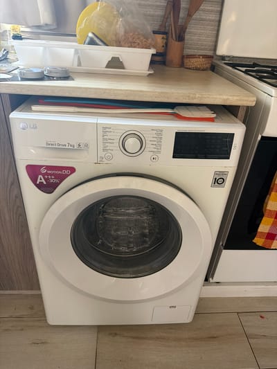 An LG front-loading washing machine is positioned on a tiled floor against a wooden panel wall. The washing machine is white with a silver door and control panel. Above the washing machine, a light-colored countertop is visible, holding various kitchen items including a white dish rack with some dishes, two round knobs, and a yellow bowl. A variety of wooden utensils are stored in containers on the countertop, and a small woven basket is also present. To the right of the washing machine, a portion of a white oven is visible. The scene suggests a domestic environment, likely a kitchen or laundry area within a residence in Dromolaxia - Meneou Municipality, Cyprus.