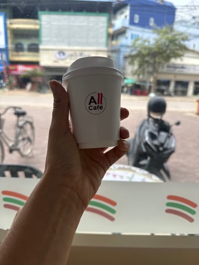 A hand holds a white disposable coffee cup with a red and black logo that reads "All Café" in front of a window. Outside the window, a blurred urban scene is visible, including buildings, a tree, a bicycle, and a motorcycle with a rider wearing a black helmet. In the foreground, below the window, a white surface with colorful stripes is visible. The image appears to be taken in Battambang, Cambodia, given the presence of the All Café chain, which is popular in the country.