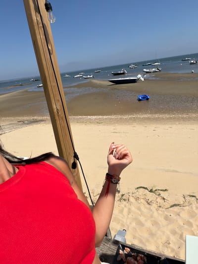 An outdoor, eye-level shot captures a beach scene with a body of water, sandy shore, and a clear blue sky. In the foreground, a person wearing a red tank top is visible from the chest up, with their arm extended to the right. The person's hand, with manicured nails, is open. They are wearing bracelets on their wrist. A wooden post with a light fixture stands to the left. In the mid-ground, several boats are anchored in the shallow water near the shore. A bright blue object, possibly a toy, rests on the wet sand. The background shows more boats in the distance, and the horizon where the water meets the sky. There is a small grill with some seafood on it in the lower right corner of the image.