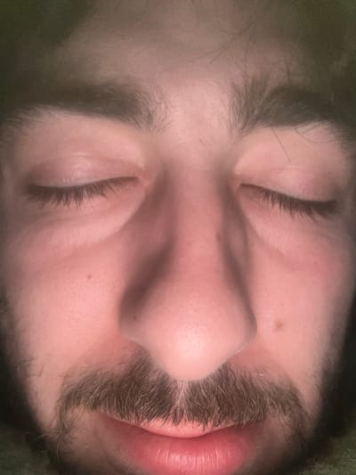 A close-up, slightly angled shot captures a person's face in repose. Their eyes are closed, and a mustache partially obscures their upper lip, which is slightly parted. The skin tone is light with visible pores and a single mole near the nose. Eyebrows are full and dark. The individual appears to be lying down, with some fabric visible at the edges, possibly clothing or bedding. The lighting is diffused, creating a soft, intimate feel.