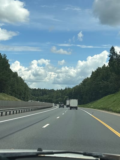 The image captures a roadside scene on a bright, sunny day in Leshkovo, Russia. The perspective is from a moving vehicle, looking forward and slightly to the right, revealing a multi-lane highway. The road is asphalt, marked with a solid yellow line separating the lanes and broken white lines indicating lane divisions. Two semi-trailer trucks are visible in the distance, traveling in the same direction.
To the left of the highway, a dense forest borders the road, with the trees forming a lush green wall. Above the treeline, a dynamic sky is filled with a mix of bright white cumulus clouds and wispy cirrus clouds against a vibrant blue backdrop. The sunlight suggests it is daytime, likely mid-morning or afternoon.
To the right of the road, a grassy embankment slopes upwards, also densely covered in green vegetation. A metal guardrail runs along the edge of the road, separating the highway from the embankment. The overall impression is one of travel on a clear, pleasant day. No people are directly visible, and no text is discernible in the image.