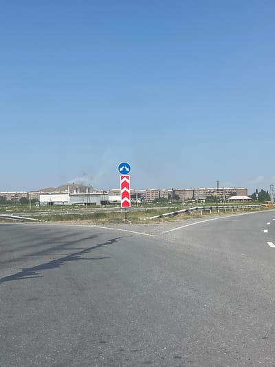 The media file depicts a roadside scene under a clear blue sky, suggesting daytime with fair weather. The perspective is from a vehicle, with the road taking up the right half of the frame, appearing to be asphalt with faint white lane markings. To the left of the road, a metal guardrail with black and white stripes separates the roadway from a grassy verge. Beyond the verge, a low-rise urban landscape is visible, characterized by various buildings, some appearing to be industrial or institutional, with one structure topped by a tall, slender tower or chimney emitting a faint plume of smoke. In the mid-ground, several residential or commercial buildings can be seen, of varying heights and architectural styles.

A prominent traffic sign is situated on a pole to the left of the road. It consists of two parts: a circular blue sign with two white arrows pointing in opposite directions, indicating a roundabout or a traffic circle. Directly below this is a rectangular sign with a red background and a series of white chevron arrows pointing to the left, signaling a curve or hazard to the left.

The overall impression is a view of a highway or major road passing through a semi-urban or industrial area. The light and shadows suggest it is mid-day. No people are directly visible, but the presence of the road and buildings implies human activity and habitation. The location context of Surenavan, Armenia, provides a specific geographical anchor to this scene.