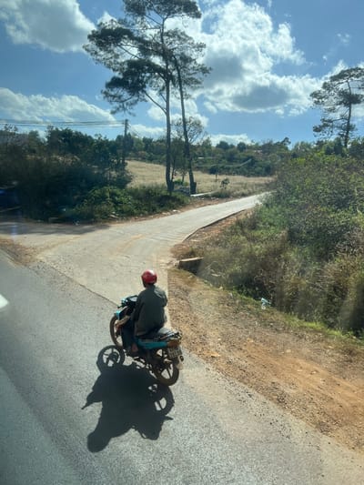 A man wearing a red helmet rides a motorcycle on a paved road. The road extends into the distance, flanked by greenery on both sides. One side has a dirt embankment and forest, while the other reveals a field. Tall trees stand to the left with a cloudy blue sky overhead. The image is slightly tilted, altering the perspective.