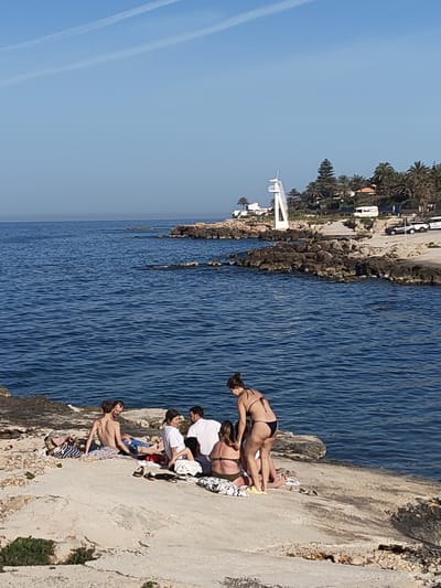 A daytime shot in Rotes, Spain, shows a group of seven people on a rocky, seaside location. To the left of the frame, two men are sitting, one shirtless and the other in blue swim trunks. In the center, four individuals are gathered, with three seated and one woman standing in a black bikini. A man is seated in a white shirt. To the right, a woman in a bikini is assisting another woman who is seated. The rocky terrain extends into the ocean. In the background, a white lifeguard tower is visible near parked cars, palm trees, and other vegetation. The sky is clear.