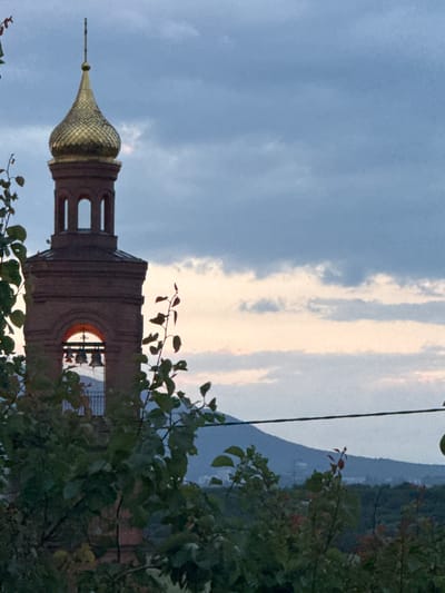The image shows a close-up view of a brick bell tower with a golden dome against a cloudy sky. The bell tower has arched openings with bells visible inside one of them. Green foliage in the foreground partially obscures the lower section of the tower. In the background, a distant mountain range is visible under a twilight sky. The scene appears to be situated in the vicinity of the city of Novoterksy, Russia, given the architectural style commonly found in the region.