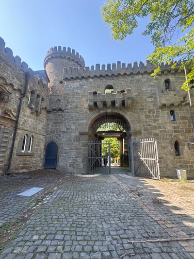 The image shows a view of the castle gatehouse of Wilhelmshöhe Palace, located in Kassel, Germany. The castle is constructed of stone blocks and features battlements, a tower, and a large arched entryway with a metal gate. The foreground is a cobblestone courtyard. Two people, partially obscured by the gate, are walking through the entryway and into the courtyard beyond. The sky is clear and blue, with the leaves of a tree visible in the upper right corner.