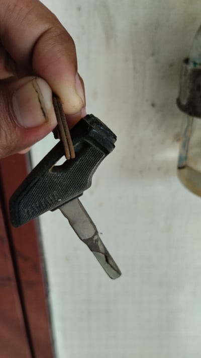 A person's hand is holding an old, black motorcycle key. The key has a textured grip and a metal blade. The background is a light-colored wall, possibly in Tanjungsari, Indonesia. There is a reddish-brown object to the left of the hand and a blurry, partially visible object to the right. The key appears worn and possibly damaged.