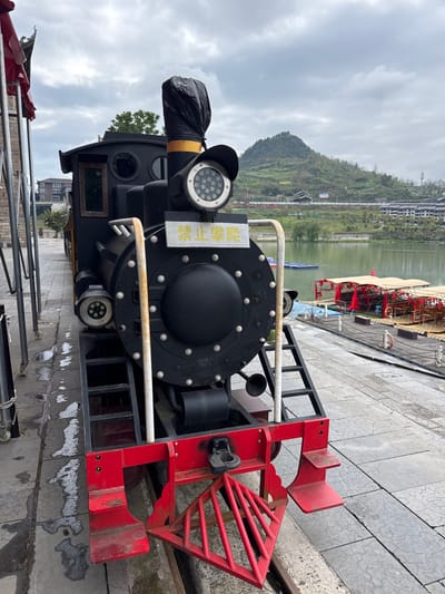 The image depicts a stationary, decorative train engine in what appears to be a recreational or tourist area, likely in Zhuoshui, China, given the presence of Chinese signage on the train. The engine is painted black with red trim and silver accents. The front of the train features a red cowcatcher and a black smokestack, partially covered with a black plastic bag. Two white handrails run along the sides of the boiler. A sign with Chinese characters is affixed to the side of the engine, close to the headlight. The ground is paved with gray stone tiles, showing water stains. In the background, a body of water reflects the overcast sky. On the opposite side of the water, a hillside is covered in greenery, with buildings nestled among the trees. Several small, brightly colored boats are docked on the waterfront behind the train. The photo is taken from a close-up perspective, focusing primarily on the front of the train engine. The overall atmosphere is somewhat gloomy due to the overcast sky.