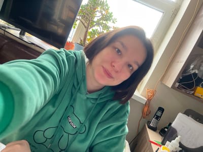 This image depicts an indoor scene in Kozlovka, Russia. The central subject is an adult female, positioned in a selfie-style shot, looking directly at the camera with a slight upward curve of the lips. She has medium-length dark hair. She is wearing a green hooded sweatshirt featuring a black graphic of a bear with the text "HSFUCK" printed vertically on its chest area.
In the background, to the left of the subject, a portion of a black flat-screen television is visible, positioned above a brown upholstered object, possibly a couch or chair. Behind the subject's head and extending to the right, a large window is present, offering a view of an overcast sky outside. On the windowsill, a potted plant with numerous green leaves is visible. Below the window, a wooden surface, likely a desk or table, holds various items. These include a coiled orange electrical cord, a black cordless telephone on its charging base, multiple sheets of paper with handwritten text, a red pen, and several small white and yellow bottles/containers. Shelves constructed from light-colored wood are visible in the upper right quadrant, containing additional miscellaneous items, some encased in clear plastic. The room is illuminated by natural light entering through the window.