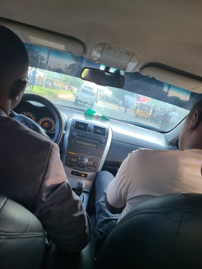 The image is taken from inside a car in Karu, Nigeria, looking forward through the windshield. The driver, a man wearing glasses and a brown suit jacket, is visible in the left foreground. To the right, a passenger wearing a light-colored t-shirt and jeans is seated. The car's dashboard and steering wheel are prominent. Outside the car, a street scene unfolds with several people walking and various vehicles, including a yellow auto-rickshaw, a white pickup truck, and motorcycles. The environment appears to be a typical urban street with buildings and trees visible in the background under a bright sky. The overall impression is a snapshot of daily life in a Nigerian city.