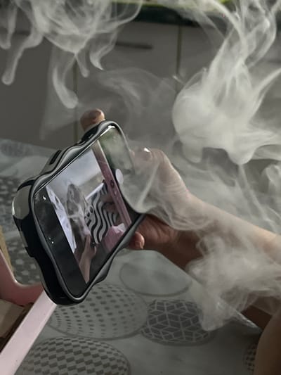 A mobile phone is being held in a hand amidst a cloud of white smoke. The phone is black and is reflecting the image of a cat on its screen. The background features a geometric pattern.
The location of this scene within Port-Katon, Russia, cannot be determined from the visual information available.