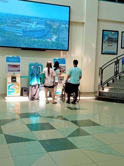 The image shows a scene inside a building in Kecamatan Margorejo, Indonesia (latitude: -6.761, longitude: 111.014). Four people are present. Three of them are standing near an automated machine, possibly a ticket machine or a payment terminal. The person farthest to the right is wearing a teal shirt and is standing beside a wheelchair. The person on the far left is wearing a blue hijab and a long gown. The person in the middle is wearing a pink shirt and black shorts. The person behind her is wearing a black shirt and dark pants. Behind the people, there is a large flat screen displaying an aerial image of a building. The building is located in front of a large grassy area. The scene is well lit and the floor is a mixture of green and white tiles. On the right side of the image, there is a staircase leading up to the next floor. The wall is white and the floor is covered in a light gray and dark gray checkerboard pattern.