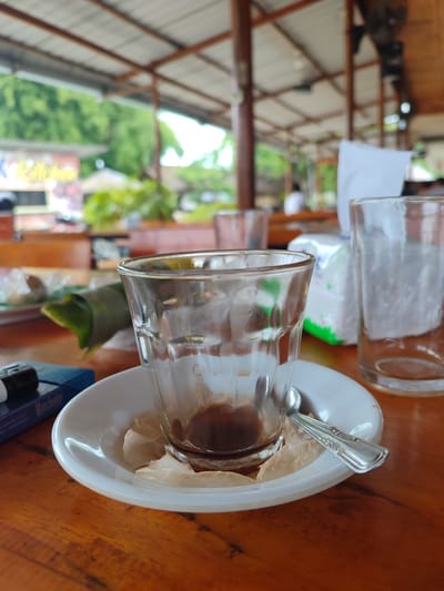 A close-up, eye-level shot shows an empty glass with a small amount of dark liquid residue at the bottom, sitting on a white saucer. A silver spoon rests on the saucer, partially under the glass. To the left of the glass is a blue cigarette pack with "Manchester" printed on it and a lighter. In the background, a second empty glass is visible, as well as a package of tissues with green accents. The scene is set on a wooden table, possibly in an outdoor cafe or dining area. The background is blurred, showing wooden beams and a roof structure, suggesting an open-air establishment. Lush green foliage is visible beyond the roof. The overall impression is that of a recently finished drink at a casual eatery. This scene could be located in Julikeude Dua, Indonesia, a region known for its natural beauty and welcoming cafes.