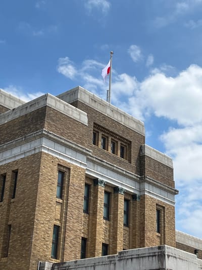 An upward-angled photograph captures the upper facade of a multi-story building, identified as being in Taito, Japan, beneath a blue sky with scattered white clouds. The building's exterior is constructed of light brown brickwork, featuring various rectangular windows with dark frames, some appearing recessed. Prominent architectural elements include multiple light grey stone or concrete cornices and horizontal banding, some exhibiting dark discoloration from weathering. Decorative brackets or capitals are visible beneath the uppermost cornices. A white Japanese flag (Hinomaru) with a central red disc is affixed to a silver flagpole on the building's roof, gently unfurling in the breeze against the sky. No individuals, animals, or specific actions are discernible within the frame.