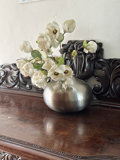 A close-up shot depicts a vase of white magnolia flowers, nestled against an ornate, dark brown, carved wooden frame. The vase is silver and egg-shaped, reflecting light. The flowers have green leaves and stems, and their petals are slightly open, revealing dark centers. The background consists of a light wall and a section of the dark frame.