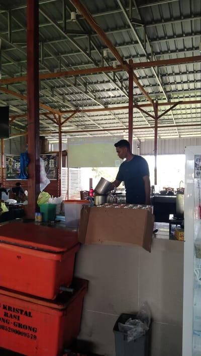 In the city of Keude Trienggadeng, Indonesia, a man is seen working behind a counter in an open-air food stall. The stall has a corrugated metal roof supported by wooden beams, with a projector screen displaying a soccer match in the background. The man, wearing a dark t-shirt, is pouring liquid from one pot to another. In front of him, stacked orange coolers are visible, with text on the lower cooler reading "KRISTAL RASI GONG-GRONG 085290009579". A green garbage can and a trash receptacle filled with plastic bags are also present. The overall scene suggests a casual, functional eatery.