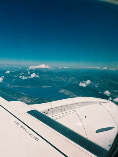 An aerial view from an airplane window shows a vast body of water, likely a bay or fjord, surrounded by mountains. The water is a deep blue, reflecting the clear sky above, which transitions from a bright turquoise near the horizon to a darker, intense blue at the zenith. A coastline with a settlement is visible along the edge of the water, featuring buildings and infrastructure. The mountains are rugged and covered in vegetation, with some parts appearing rocky. Clouds are scattered across the sky, casting shadows on the landscape below and adding depth to the scene. In the foreground, a portion of an airplane wing is visible, painted white with a prominent dark stripe. Black lettering on the wing reads "DO NOT STEP OUT OF THIS AREA." The engine housing of the aircraft is also partially visible, a complex white structure with various panels and details. The scene suggests a flight over a coastal mountainous region, possibly the Bay of Kotor in Montenegro, with the city of Kotor or a similar settlement nestled along the water's edge. There are no people or animals visible in the image. The overall impression is one of serene aerial travel over a beautiful, natural landscape.