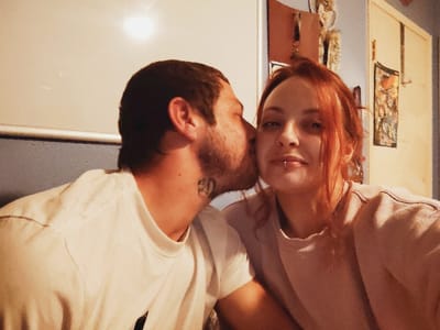 A man and woman pose for a selfie indoors. The man, on the left, wears a white t-shirt and has a beard. He is kissing the woman's cheek. The woman wears a light pink shirt and has red hair and a lip piercing. Behind them is a white wall, a blue wall, a cabinet, and artwork. The lighting is soft.