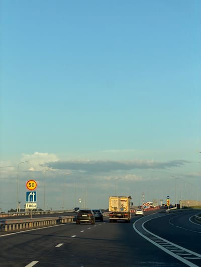 The image depicts a highway in Zimnyaya Gorka, Russia, under a clear blue sky with scattered clouds. On the left side of the road, traffic signs indicate a speed limit of 50, and lanes for straight ahead and turning right. Several vehicles, including cars and a truck, are visible on the multi-lane highway. Streetlights line the road, and barriers separate the lanes. The environment appears to be a sunny day, possibly late afternoon, given the angle of the light casting shadows. The road surface is asphalt with white lane markings.