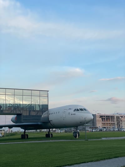 The media file is an image of an airplane on a tarmac at an airport. The airplane, identified by the text "ЯК-42Д" on its fuselage, is white with several windows visible on its front. Its landing gear is deployed, suggesting it is either on the ground or preparing for takeoff/landing. Adjacent to the airplane is a modern structure with a glass facade, reflecting the sky and clouds. Beyond this structure, a long stretch of vibrant green grass lines a paved walkway. In the background, there are airport buildings under construction, with scaffolding and incomplete structures. Several vehicles, including cars and what appear to be airport service vehicles, are parked in front of the buildings. The sky is a clear blue with some scattered white clouds, indicating fair weather. The lighting suggests it is either morning or late afternoon. The overall scene depicts a functional airport environment with ongoing development. No people are visible in the frame.
