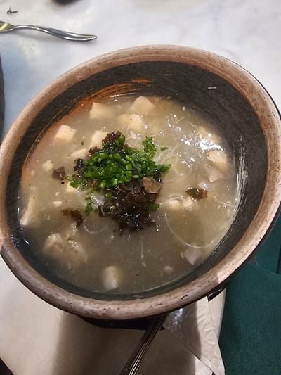 A top-down shot showcases a bowl of soup, likely Asian-inspired, featuring a creamy, pale broth. The soup contains soft, white tofu cubes, translucent noodles, and is garnished with a cluster of chopped green herbs and dark, possibly dried, mushrooms. The bowl is crafted from a dark, rustic material with a tan rim. A fork rests on a napkin next to the bowl.