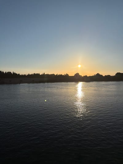 This is a landscape photograph taken during sunset in Arles, France. The image depicts a serene body of water, likely the Rhône River, with a distinct reflection of the sun shimmering on its surface. The sun itself is visible, a bright orb just above the horizon, casting a warm glow across the sky. A silhouetted treeline with sparse buildings runs along the far bank of the river, creating a natural border between the water and the sky. The sky transitions from a soft blue near the top to a pale yellow near the horizon, characteristic of a sunset. The water appears calm, with subtle ripples creating texture. The overall mood of the image is peaceful and tranquil, evoking the quiet beauty of a day's end in a picturesque French setting. No text is visible in the image.