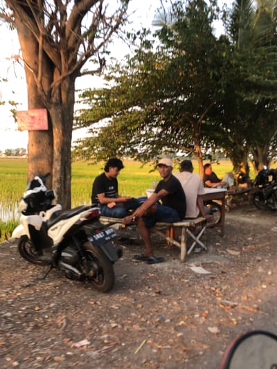 In the city of Keboguyang, Indonesia, on a dirt road bordering a rice paddy, several men are gathered under a large tree. A white scooter is parked on the left, its license plate reading "W 5887 NOH". A group of men are seated on a low bamboo table and benches. One man, wearing a black t-shirt and blue jeans, is facing the camera. Another man in a white t-shirt and blue jeans is seated with his back to the camera, while a third man, in a black t-shirt, sits further away. In the background, a rice field stretches out, and other trees dot the landscape. The sun is low in the sky, casting long shadows and creating a warm, golden light.