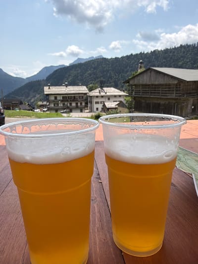 Two plastic cups filled with golden-hued beer and topped with white foam sit on a wooden table in the foreground. In the background, traditional houses with stone and wooden elements are nestled amidst lush green hills and mountains. The sky above is a mix of blue with scattered white clouds. The scene appears to be a scenic viewpoint in Sauris / Zahre, Italy, known for its unique Germanic dialect and alpine architecture.