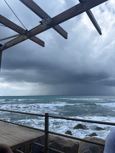 The image displays a coastal scene in Episkopi Lemesou, United Kingdom, under a heavily overcast and stormy sky.
In the upper left quadrant, a wooden pergola structure is visible, composed of dark brown exposed beams. Mounted on these beams are two white dome-shaped security cameras (CCTV) and a single exposed incandescent-style light bulb hanging by a black wire.
Below, in the lower left, a section of a light brown wooden table or deck is visible, with a person's shoulder in a white shirt partially seen on the extreme lower right. A dark grey metal railing runs horizontally across the lower portion of the frame, separating the wooden area from the water. Large, irregular grey rocks form a breakwater or shoreline directly beneath the railing.
The midground is dominated by the turbulent turquoise-blue sea, characterized by numerous white-capped waves breaking and churning towards the shore. In the background, the sky is uniformly dark grey with heavy storm clouds. A distinct vertical band of falling rain is visible over the distant sea, indicating active precipitation.