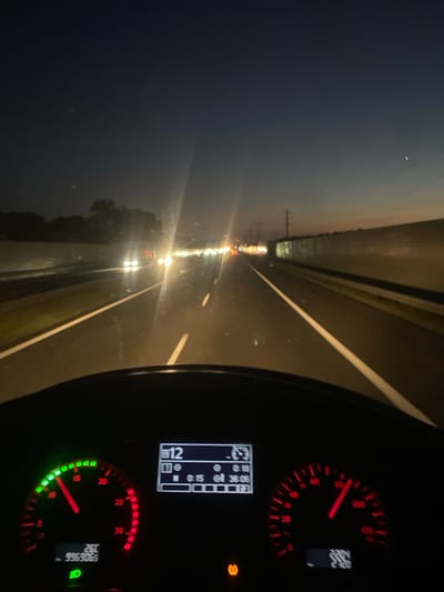 The image captures a view from the driver's perspective within a vehicle, likely a truck, on a highway at dusk or early night. The road ahead is illuminated by oncoming headlights and streetlights, creating streaks of light due to the camera's exposure. The dashboard displays various indicators, including a speedometer showing a speed between 20 and 25, a digital display showing "12" and some time readings, and another speedometer. The environment suggests a rural or suburban setting outside of a major city. The sky is a deep blue, transitioning to orange near the horizon, and a faint star or planet is visible. There are trees and vegetation visible on the left side of the road, and a sound barrier or wall lines the right side. The image was taken on the A4 highway near Kokotów, Poland, a location known for its agricultural and rural landscape.