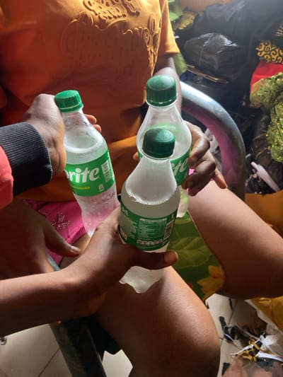 Two people holding Sprite bottles are seated next to each other. The bottles appear to be cold, with condensation forming on the outside. One person is wearing a gold-colored shirt or dress, while the other has on a black and red sleeve. The people are seated in metal chairs, with the legs of the person on the right visible. In the background, there is what appears to be clothing and other items.