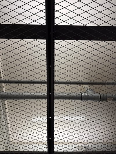 A vertical black metal bar bisects the image, a diamond-patterned metal mesh lies in front of a light-colored wall, and a galvanized metal pipe runs horizontally across the mesh. The pipe is bent into a T-junction with two sections of black tape wrapped around it at the junction. The lighting is bright and casts shadows from the mesh onto the wall. The scene is located on a street in Duisburg, Germany.
