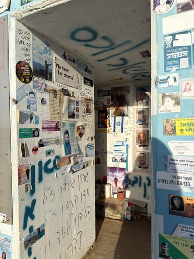 The image captures the interior of a concrete, box-like structure, likely a public shelter or memorial shrine, seen from above. The walls are covered in a dense mosaic of photos, handwritten notes, printed flyers, and graffiti, predominantly in Hebrew.
The top edge of the structure is painted light blue, while the inner walls are white. Sunlight streams in, creating strong shadows across the back and right walls.
**Details on the Walls:**
* **Top Blue Strip:** Features various flyers and photos. One prominent blue flyer includes "1223" and a QR code. Several individual photos are visible, including one identified by OCR as "שני גבאי" (Shani Gabay), a victim of the 2023 Nova Festival attack, along with the text "לעולם לא נוכל לעצבות" (We will never surrender to sadness) and "לנצח בליבנו" (Forever in our hearts). Other flyers are in green and white.
* **Right Inner Wall:** Displays four rectangular portraits of young men, possibly soldiers or victims, arranged horizontally. Below these are numerous handwritten messages in Hebrew and a small Israeli flag. Towards the bottom, a clear glass container and a small bottle of "AJAX" are visible on the ground level. More papers and handwritten notes fill this wall.
* **Bottom Inner Wall:** Is extensively covered. A large image with the English text "No time for drama" is prominent. Another photo shows a person with outstretched arms against a cloudy sky. Many smaller individual photos, newspaper clippings, and personal messages (some including hearts and Stars of David) are affixed. A circular sticker with Hebrew text and a graphic is also present. A photo of a figure resembling Jesus in blue robes can be seen, along with a Batman logo sticker. Handwritten messages and blue spray-painted graffiti are scattered throughout.
* **Left Inner Wall:** Features large blue spray-painted Hebrew graffiti, reading "סולי" (Soli) or "סוד" (Sod), alongside more handwritten messages and various photos and papers.
**Visible Text and Themes:**
The texts extracted by OCR and visually identified include:
* "שני גבאי" (Shani Gabay)
* "לנצח בליבנו" (Forever in our hearts)
* "הקושי הוא זמני הגאווה היא נצחית" (Difficulty is temporary, pride is eternal)
* "תהיה האור שאתה רוצה לראות באחרים" (Be the light you wish to see in others)
* "No time for drama"
* "1223"
* "MDY", "SUN DON'T"
* "אני חי" (I am alive)
* "התחלתם משהו תסיימו" (You started something, finish it)
* "לדוד מלך ישראל" (To David King of Israel)
The accumulation of personal photos, memorial messages, and specific names, particularly in the context of recent events in Israel, suggests this structure serves as a spontaneous memorial or shrine dedicated to victims of conflict or tragedy. The overall atmosphere conveys remembrance, grief, but also resilience and hope.