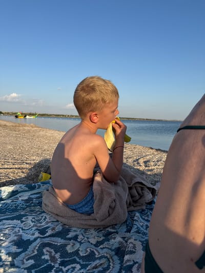A young boy with blond hair is sitting on a towel at the beach, eating a banana. He is wearing blue swim trunks. The boy is shirtless and appears to be enjoying his snack. Behind him, the calm water of the sea stretches out to the horizon under a clear blue sky. The beach is covered in pebbles. In the distance, there are a few small boats on the water. The sun is low in the sky, casting a warm glow and long shadows, indicating it is likely late afternoon or early evening. A person's legs, wearing a green swimsuit, are visible in the upper right corner of the frame, suggesting they are sitting next to the boy. The overall mood is relaxed and peaceful, capturing a moment of simple enjoyment at the seaside.
