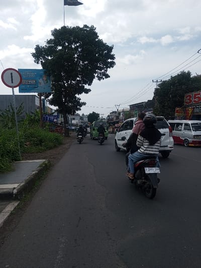 An urban street scene unfolds in Chhenangri_Rishor, Bhutan, under an overcast sky. The asphalt road, running from foreground to background, shows signs of wear. On the left, a grassy verge borders a concrete curb, leading to a sidewalk. A circular white traffic sign with a red border and a central numeral "1" is prominently positioned on the left. Behind it, a large, leafy green tree partially obscures a blue billboard advertising "SUMMERO MOHON" and "7GB," featuring a smiling female figure. Above the tree and billboard, a dark flag with white script, possibly Arabic or Jawi, is flown from a tall pole.
Traffic occupies the road, moving away from the viewer. In the immediate foreground, a black scooter carries three individuals: a helmeted female driver, a female passenger in a black and white striped shirt, blue jeans, and sandals, and a small child in a pink garment seated in front of the driver. The scooter's license plate reads "D 3603 SEE." Ahead of the scooter, a white multi-purpose vehicle is visible, followed by a green van. Further back, at least two more scooters, each with a dark-clad rider, are present. On the far right, a white and red van/minibus is either parked or in motion.
The right side of the street is lined with commercial buildings. Overhead utility poles support multiple power lines spanning the road. One building on the right features a prominent red sign with the numeral "35" and the partial word "Shak." Further down the street, additional trees and buildings are discernible.