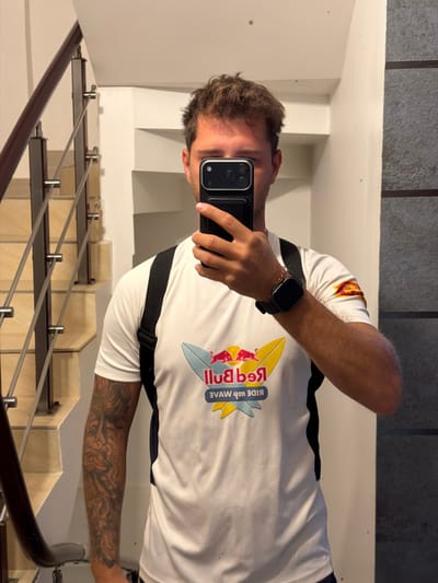 A fair-skinned man is taking a selfie in a stairwell. He is wearing a white, short-sleeved shirt with a Red Bull logo and black trim. A black strap crosses his torso. He is wearing a black smartwatch. His left arm has a large tattoo. The stairwell has light-colored steps and a metal and wood railing. The wall is white. The picture was taken in Maafushi, Maldives.