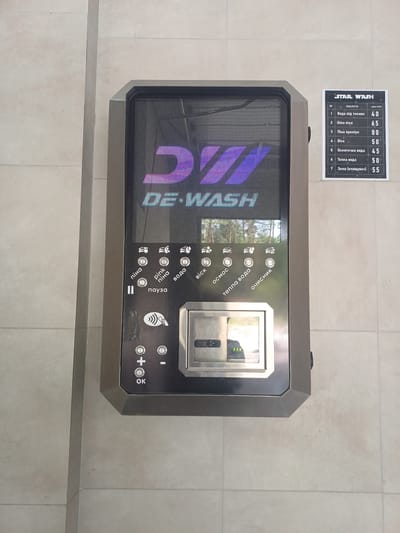 The image shows a self-service car wash machine with a digital display and control panel. The machine is dark gray with illuminated blue and purple graphics. A price list board, titled "STAR WASH," is affixed to the wall to the right of the machine. The price list details services and their costs in Ukrainian Hryvnia (UAH). The machine has various buttons with icons and Ukrainian labels, including "піна" (foam), "pink піна" (pink foam), "вода" (water), "віск" (wax), "осмос" (osmosis), "тепла вода" (warm water), "очисник" (cleaner), and "пауза" (pause). There is also a card reader and buttons for adjusting time or price (+/-) and an "OK" button. The machine is mounted on a light-colored tiled wall. The environment appears to be outdoors, with the reflection of trees visible on the machine's display. Based on the presence of the "STAR WASH" brand and typical self-service car wash locations, this machine is likely located in the city of Obukhiv, Ukraine. There are no people visible in the image.