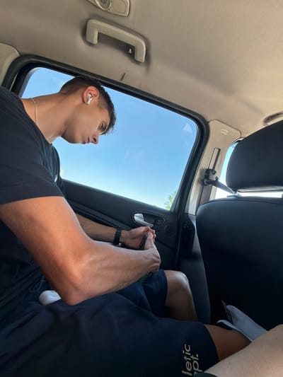The media file depicts a young man inside a vehicle, possibly a car. He is wearing a black t-shirt and appears to be looking out of a sunroof or window, with the bright blue sky and some greenery visible outside. He has an earbud in his left ear and a thin necklace around his neck. His right arm is extended, and he is holding a pen or stylus in his hand, seemingly interacting with something just out of frame. The interior of the car is visible, including parts of the headliner and a handle. The overall lighting suggests it is daytime, likely sunny. The man's expression is neutral, focused on whatever he is looking at or interacting with. There is visible text on the man's shorts, though it is partially obscured and inverted, reading "letic.ept." which could be a fragment of a brand name. The scene implies a moment of rest or transit.