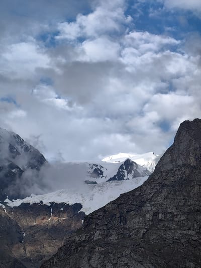 The image depicts a mountainous landscape with a glacier and snow-capped peaks under a cloudy sky. The foreground is dominated by a rugged, dark-colored rock formation on the right side, with visible texture and layering. To the left, a steep, rocky slope is partially obscured by mist and clouds, revealing patches of snow and ice. Further in the background, a large glacier extends across the frame, its surface appearing smooth and undulating, with crevasses visible. Behind the glacier, sharp, snow-covered mountain peaks pierce through the clouds, reflecting the ambient light. The sky is filled with a mixture of white and grey clouds, with patches of blue sky showing through. There are no people or man-made objects visible in the scene. The overall impression is one of a remote and majestic natural environment.