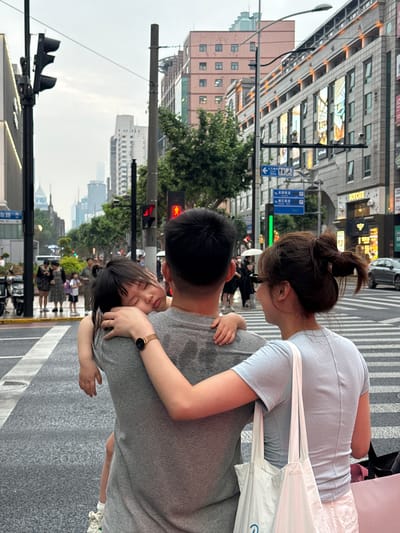 An urban street scene, rotated 90 degrees counter-clockwise, depicts a family on what appears to be a wet road or sidewalk, suggesting recent rain. In the foreground, a man in a grey t-shirt is seen from behind/side, carrying a young child who is nestled and seemingly asleep or resting on his shoulder. The child has dark hair and a small hand is visible. Next to them, a woman, also seen from behind/side, wears a light blue t-shirt, white shorts, and has dark hair tied in a bun. She carries a white shoulder bag on her left side and wears a watch with a gold strap and dark face on her left wrist.
The setting is a bustling city street, identified as Xuhui District, China. In the background, tall, multi-story buildings line the street, some with illuminated windows or signs. One prominent building sign in the mid-ground clearly reads "BOYCAFE" in English, alongside Chinese characters. Traffic infrastructure includes zebra-striped crosswalks, traffic lights (one showing red), street lamps, and blue road signs with white Chinese characters and symbols, including a wheelchair accessibility symbol and "100m." Other pedestrians are visible in the blurred background. The sky is overcast, and the overall lighting suggests either a heavily cloudy day or late afternoon/early evening. The image captures a tender, candid moment of parental care amidst the urban environment.
