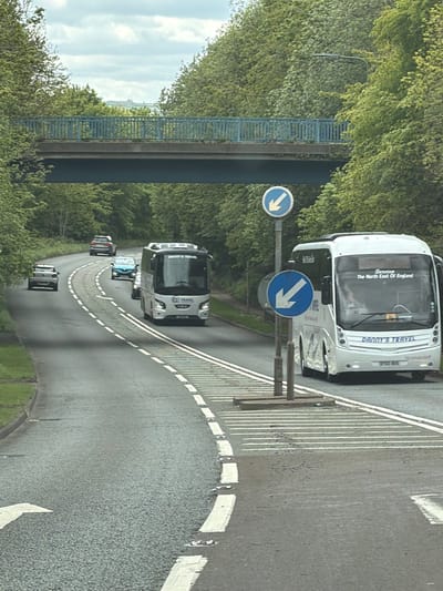 The image shows a road scene in Witton Gilbert, United Kingdom. The road curves to the left and is lined with trees on both sides. A blue pedestrian bridge spans the road in the background. Traffic is flowing in both directions, including a white coach bus with "Danny's Travel" and "The North East Of England" branding, followed by other vehicles. Road signs with blue arrows indicate the direction of travel. The road surface appears wet or slightly damp.