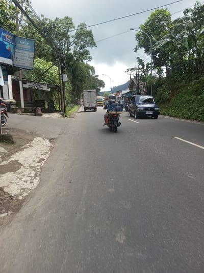 This image depicts a daytime view of a multi-lane asphalt road in the city of Ura-Dozhi, Bhutan, under an overcast sky. The road features a white solid line on its right edge and a white dashed center line, indicating two-way traffic. A person on a black motorcycle, viewed from the rear, is centrally positioned in the frame, traveling away from the viewer. The rider wears a dark helmet and dark clothing. A blue plastic crate containing various items is secured to the rear rack of the motorcycle.
Ahead of the motorcycle, a light-colored box truck and a yellow truck are traveling in the same direction. In the opposing lane, a dark-colored van, possibly a Toyota Kijang, with a partially visible license plate (beginning with 'D' or 'P'), is approaching.
To the immediate left of the road, there is a concrete/asphalt shoulder. Further left, a small, dark-roofed shelter structure, possibly a bus stop, is visible, with a sign indicating "HALTE CANDIWULAN" and other text. Above this structure, a billboard displays "TOP UP" and logos for digital payment services, including OVO, GoPay, and Grab. Utility poles with numerous overhead wires are present. Dense green foliage and mature trees line the left side of the road.
To the right of the road, a steep embankment is covered in thick green vegetation and trees, including banana plants. A streetlight pole extends over the right lane. In the distance, several buildings and structures are partially visible through the trees, suggesting a semi-urban environment. The scene indicates moderate vehicular traffic amidst lush greenery.