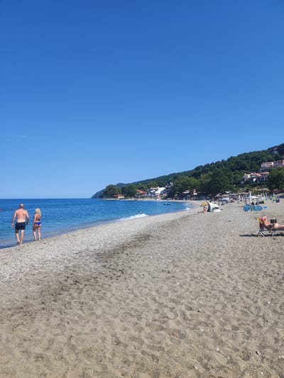 The image shows a beach in the Municipal Unit of Melivia, Greece. In the foreground, a man and a woman are walking on the sandy beach, approaching the water. The man is wearing dark swim trunks, and the woman is wearing a blue bikini. The beach has a transition from sand to pebbles closer to the water. In the background, the turquoise sea meets the shore, and beyond that, a tree-covered hillside is dotted with white buildings. Further down the beach, several people are seen relaxing under umbrellas, sunbathing on lounge chairs, or swimming. The sky is clear and bright blue.