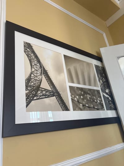 The image shows a framed collage of black and white photographs hanging on a yellow wall with white molding. The collage features an image of a large metal structure, possibly a bridge or tower, in the upper left. To the right of this, there are three smaller images: one of draped fabric, another of a branch with buds, and a third of what appears to be dense foliage. To the right of the collage, a white door is partially visible. The image is taken from a slightly elevated angle. There are no people or animals visible in the image. The context of San Francisco, USA, is not evident from the visual information provided.